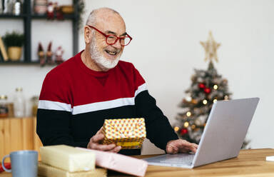 Happy senior man wearing eyeglasses holding gift box using laptop at home - GIOF15367