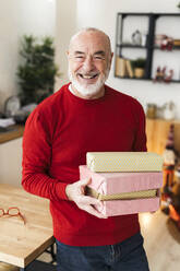 Cheerful senior man holding gift boxes standing by table at home - GIOF15370