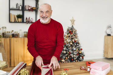 Fröhlicher älterer Mann beim Einpacken von Weihnachtsgeschenken auf dem Tisch zu Hause - GIOF15380