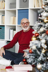 Happy senior man looking at Christmas tree sitting in front of bookshelf - GIOF15391