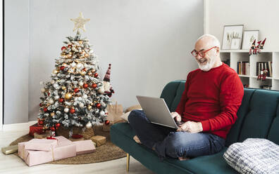 Lächelnder älterer Mann mit Laptop auf dem Sofa im Wohnzimmer zu Hause - GIOF15396