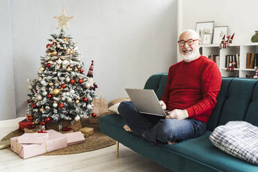 Lächelnder älterer Mann mit Laptop, der zu Hause auf dem Sofa sitzt - GIOF15397