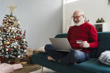 Happy senior man holding credit card doing online shopping sitting on sofa at home - GIOF15398