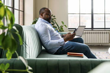 Businessman holding laptop using mobile phone sitting on sofa in office - VPIF05603