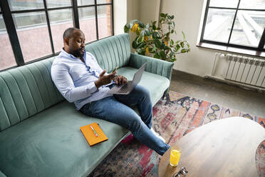 Businessman using smart phone sitting with laptop on sofa in office - VPIF05607