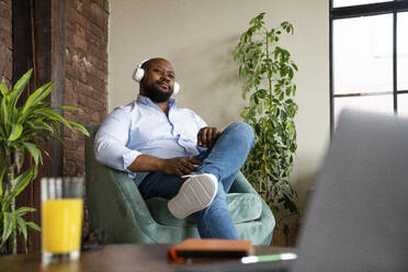 Geschäftsmann hört Musik über drahtlose Kopfhörer auf einem Sessel im Büro sitzend - VPIF05613