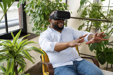 Smiling businessman wearing virtual reality simulator sitting on chair in office - VPIF05615
