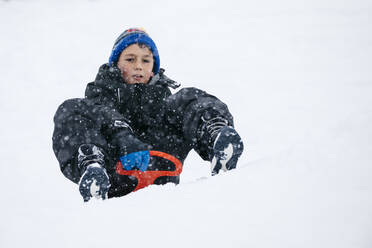 Boy wearing knit hat tobogganing on snow in winter - JRFF05359