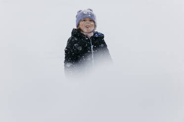 Mädchen mit Strickmütze, die die Zunge herausstreckt, steht im Schnee - JRFF05363