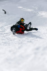 Playful boy wearing ski goggles tobogganing through snow - JRFF05366