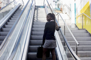 Smiling young woman with laptop bag standing on escalator - RFTF00189