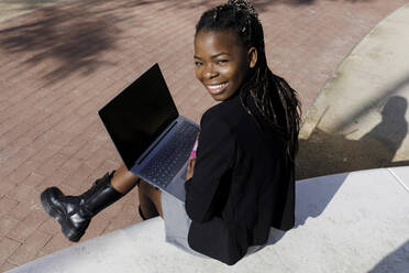 Glückliche junge Frau mit Laptop auf einem Betonsitz sitzend - RFTF00192