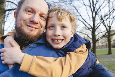 Smiling boy embracing father in public park - IHF00773