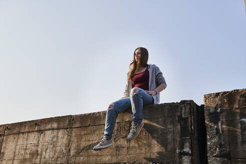 Junge Frau sitzt auf einer Betonmauer und schaut in die Ferne - MMIF00282