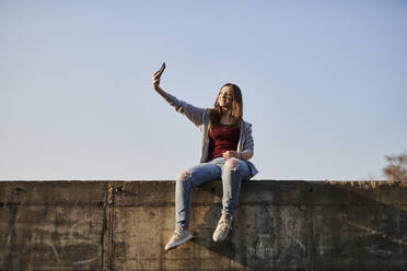 Junge Frau sitzt an einer Betonwand und macht ein Selfie - MMIF00286