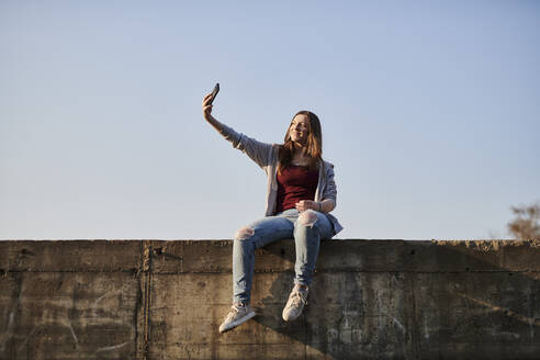 Young woman sitting on concrete wall taking a selfie - MMIF00286