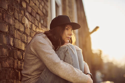 Waiting young woman with baseball cap sitting on the ground hugging knees - MMIF00300