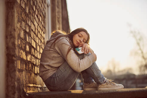 Young woman hugging knees enjoying the evening sun - MMIF00304