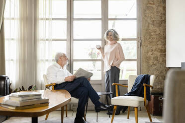 Smiling senior woman holding coffee cup talking with man in living room at home - EIF03663