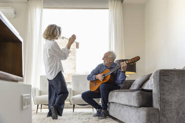 Senior man playing guitar looking at woman dancing in living room at home - EIF03671
