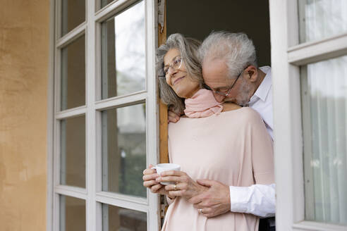 Zärtlicher älterer Mann, der eine Frau umarmt, die einen Kaffee hält, während er am Fenster steht - EIF03679
