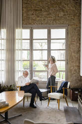 Frau mit Kaffeetasse, stehend mit Hand in der Tasche, neben einem Mann, der im Wohnzimmer zu Hause Zeitung liest - EIF03683