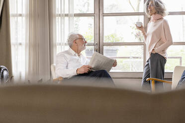 Smiling woman holding coffee cup talking with man holding newspaper in living room at home - EIF03684