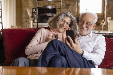 Happy senior couple having video call on mobile phone sitting on sofa at boutique hotel - EIF03694