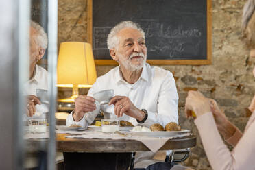 Senior man holding coffee cup sitting with woman at breakfast table in boutique hotel - EIF03709