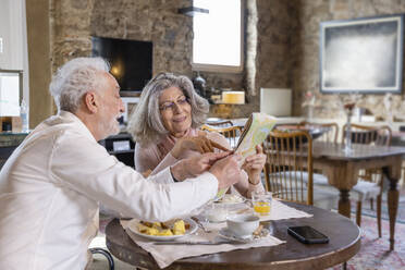 Ein älterer Mann zeigt einer Frau, die am Frühstückstisch in einem Boutique-Hotel sitzt, einen Stadtplan - EIF03713