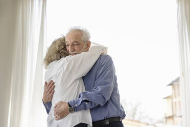 Älteres Paar, das sich umarmt und zu Hause am Fenster steht - EIF03715