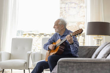 Senior man playing guitar sitting on sofa in living room at home - EIF03718