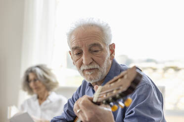 Älterer Mann spielt Gitarre mit Frau im Hintergrund zu Hause - EIF03719