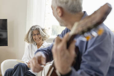 Happy woman looking at man playing guitar at home - EIF03720