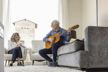 Senior woman looking at man playing classical guitar sitting on sofa at home - EIF03721