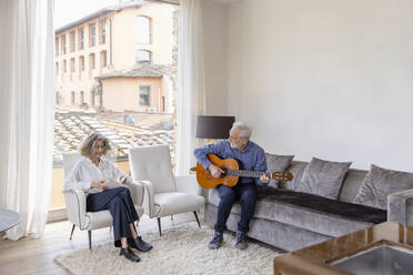 Senior man playing guitar looking at woman reading book in living room - EIF03722