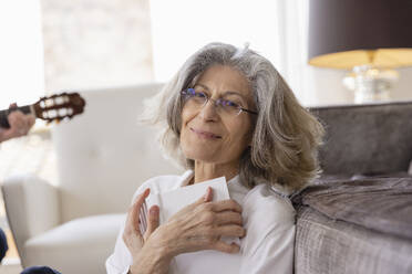 Smiling senior woman wearing eyeglasses sitting with book by sofa at home - EIF03724