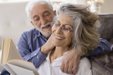 Senior man embracing woman reading book in living room at home - EIF03725