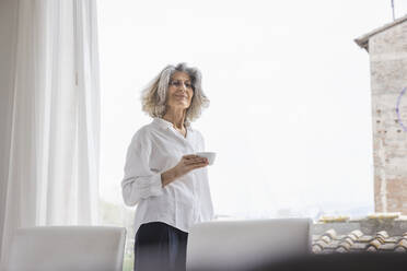Lächelnde Frau mit Kaffeetasse vor einem hellen Fenster zu Hause - EIF03736