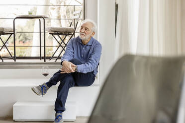 Älterer Mann sitzt mit übereinander geschlagenen Beinen vor einem Fenster zu Hause - EIF03738
