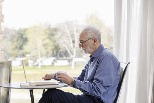 Älterer Mann mit Laptop sitzt am Tisch in einem Hotelappartement - EIF03748