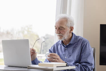Senior man with laptop and coffee cup sitting by window at hotel apartment - EIF03749