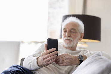 Senior man using smart phone sitting on sofa at hotel apartment - EIF03751