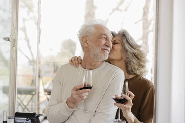 Senior woman holding wineglass kissing man in front of window at hotel apartment - EIF03753