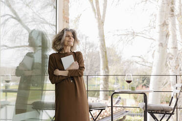 Lächelnde ältere Frau mit Buch, die sich an ein Glasfenster auf einem Balkon lehnt - EIF03760