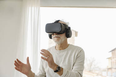 Senior man wearing virtual reality simulator gesturing standing in front of window - EIF03762
