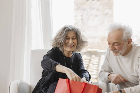 Happy senior couple with shopping bags sitting by window at boutique hotel - EIF03766