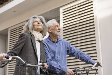 Happy senior couple standing with bicycles in front of building - EIF03778