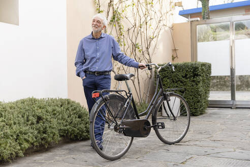 Älterer Mann mit Hand in der Tasche, der neben einem Fahrrad auf dem Gehweg steht - EIF03780