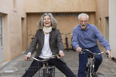Happy senior couple riding bicycles outside building - EIF03783